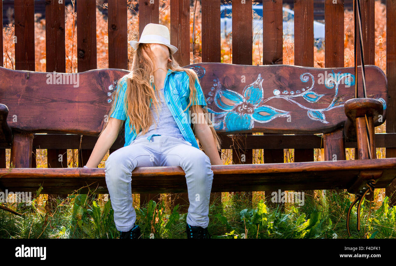 Blond, 11 Jahre altes Mädchen im blauen Hemd sitzt auf einer Hollywoodschaukel, bedeckte ihr Gesicht mit einem weißen Hut. Herbstfarben Stockfoto