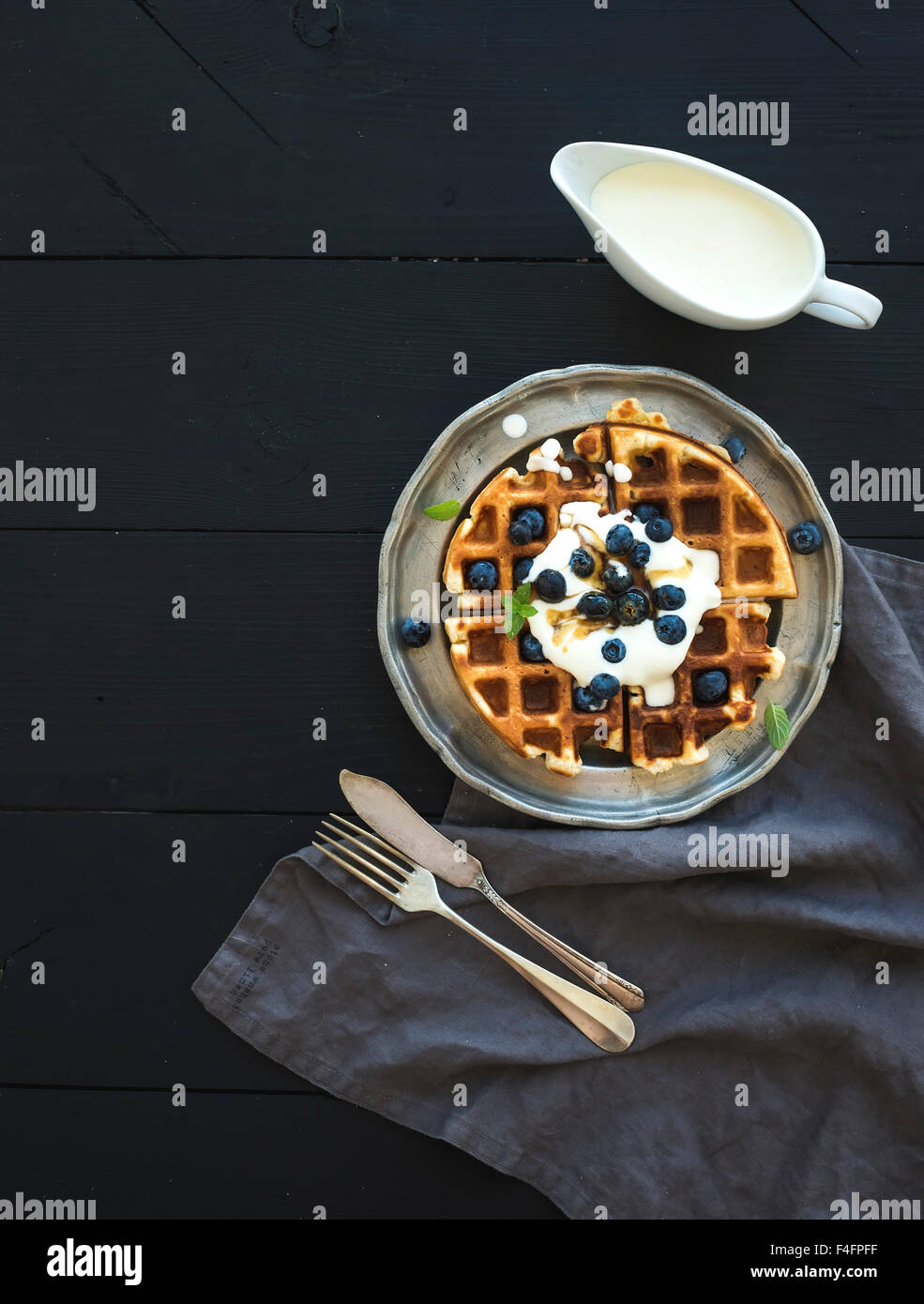 Weiche belgische Waffeln mit Heidelbeeren, Honig und Schlagsahne auf Vintage Metallplatte über schwarzem Holz, Draufsicht, co Stockfoto