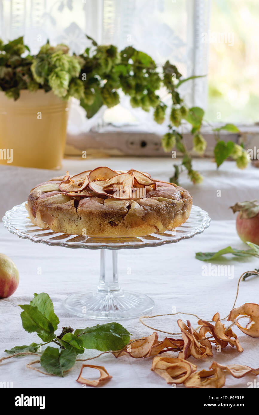 Hausgemachter Apfelkuchen auf Glas mit gebackenem, frischen und getrockneten Äpfeln über Tisch mit weißer Tischdecke stehen. Mit rustikalen Fenster im bac Stockfoto
