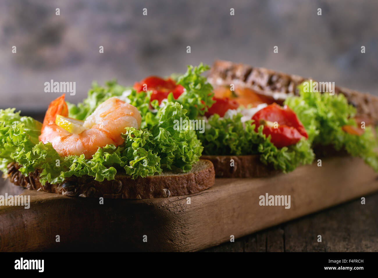 Nahaufnahme eines Sandwiches mit Vollkornbrot, frischem Salat, Feta-Käse, Tomaten, Garnelen und gesalzener Lachs auf Holz schneiden Bo Stockfoto