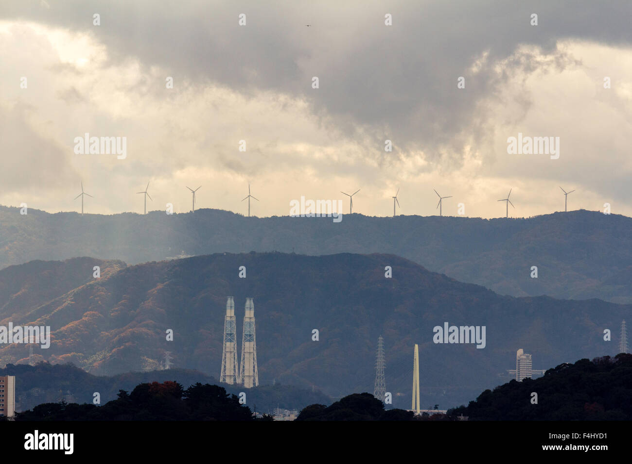 Wakayama, Japan, silhouetted Windenergieanlagen auf die Berge in der Ferne mit verschiedenen Industrieschornsteinen im Vordergrund. Regen, Sturm, aber einige Sonnenlicht. Stockfoto