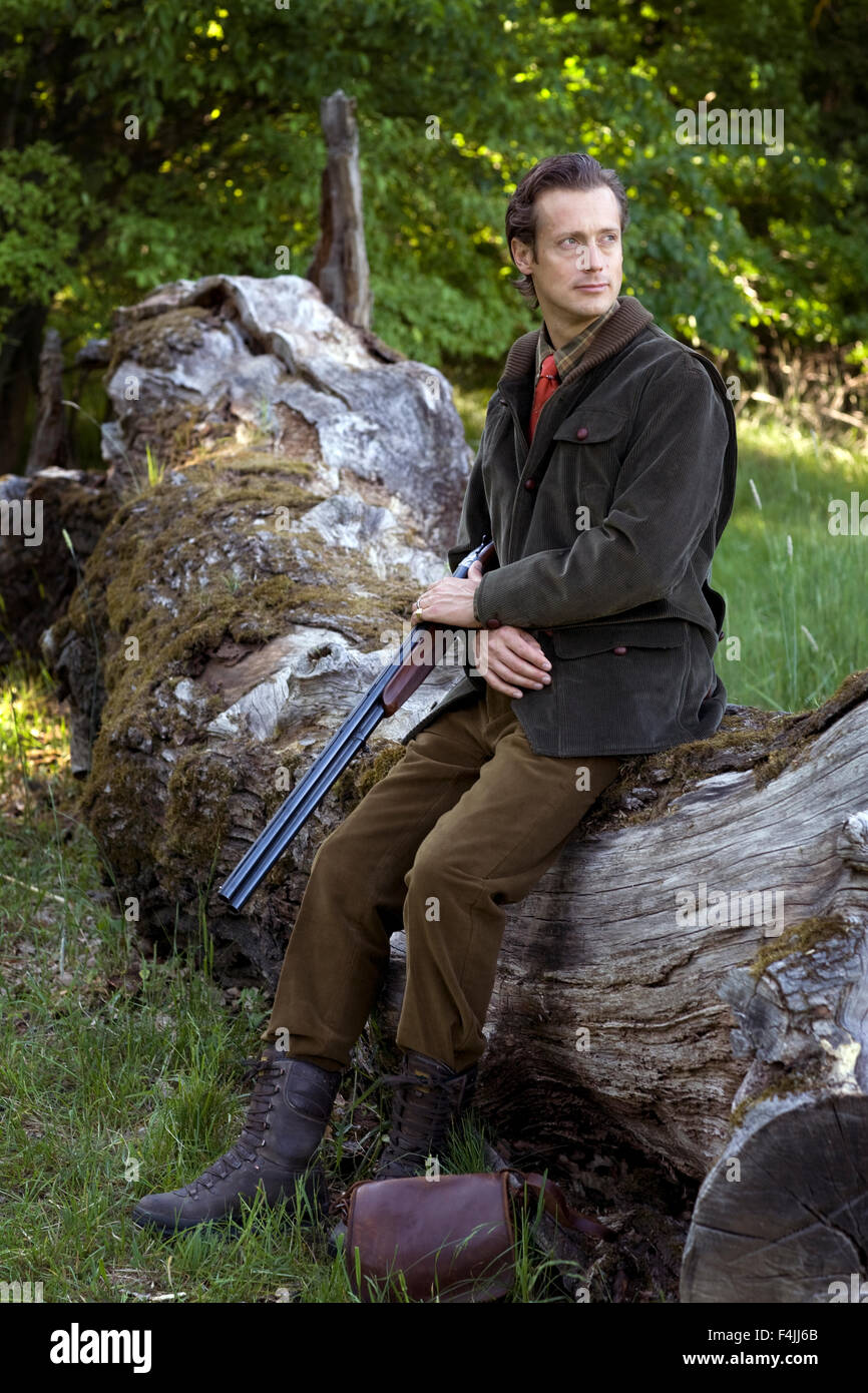 Mitte erwachsener Mann sitzend auf Baumstamm, hält Gewehr und wegsehen Stockfoto