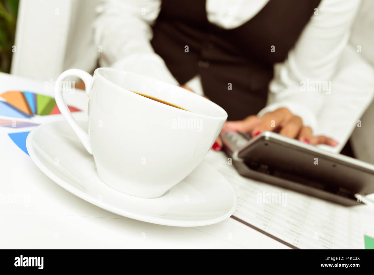 eine Geschäftsfrau, die mit einer elektronischen Rechner in ihrem Büro mit einem Schreibtisch voller Diagramme und eine Tasse Kaffee Stockfoto