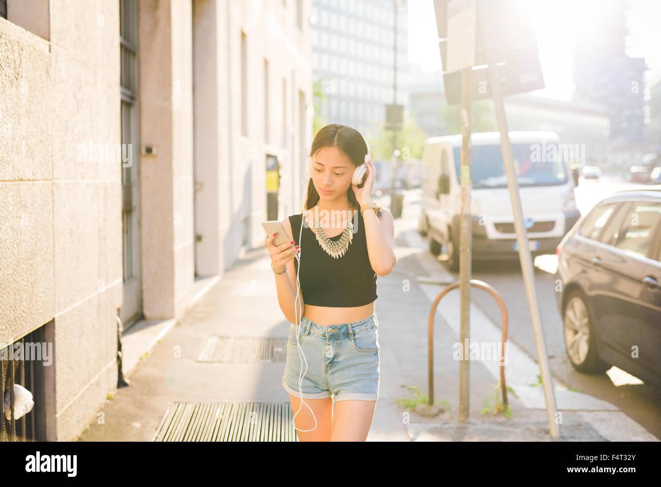 Knie-Figur des jungen schönen asiatischen Frau Wandern im Freien in der Stadt, hören Musik mit Kopfhörer, während mit einem Smartphone auf der Suche nach unten die Bildschirm - Technik, Musik, entspannende Konzept Stockfoto