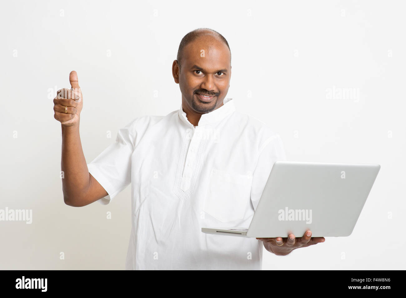 Porträt der Reife lässig indischen Geschäftsmann mit Laptop-Computer, an Kamera und Daumen nachschlagen, stehend auf einfache Zeitmessung Stockfoto