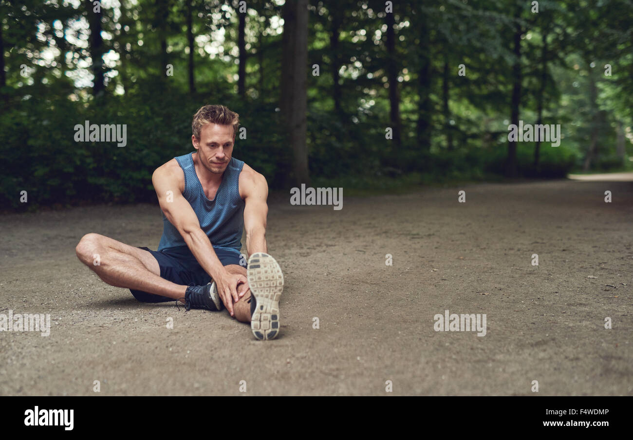 Schöner Junge männliche Sportler Lockerungsübungen vor dem Training sitzen auf dem Boden in einem bewaldeten Park tun stretching Übungen einer Stockfoto