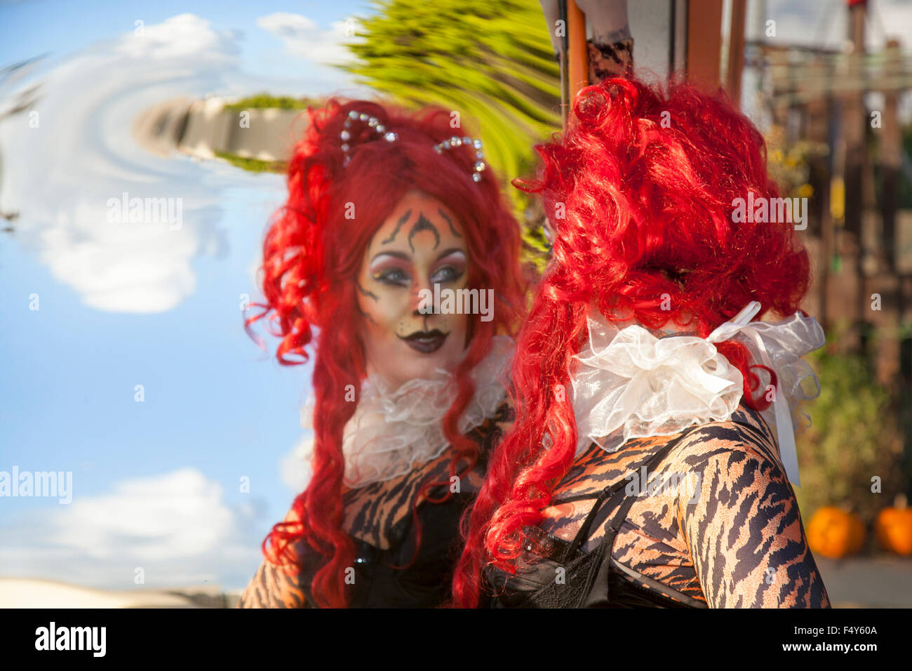 Frau mit roten Haaren Perücke, in Cat Suit mit Spaß und interessante wider Bilder zur Verzerrung des konvexen und konkaven Spiegel geben sich wiederholende Überlegungen; eine optische Illusion von Gesichtern in einem funhouse Spiegel. Das lustige Gesicht verzerrte person Reflexionen über das Thema mit einem gebogenen Spiegel ist eine traditionelle Quelle der Unterhaltung im Messegelände Halle der Spiegel. Stockfoto