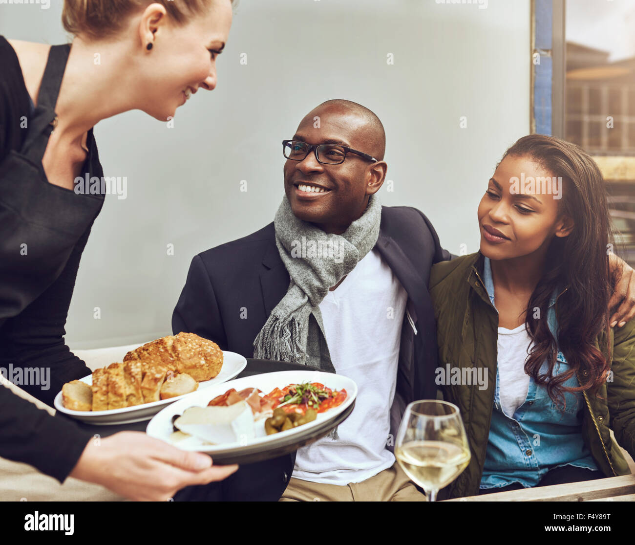 Romantische junge afrikanische amerikanische paar sitzen Arm in Arm sein Abendessen von einer Kellnerin serviert, wie sie an einem Tisch im Restaurant sitzen Stockfoto