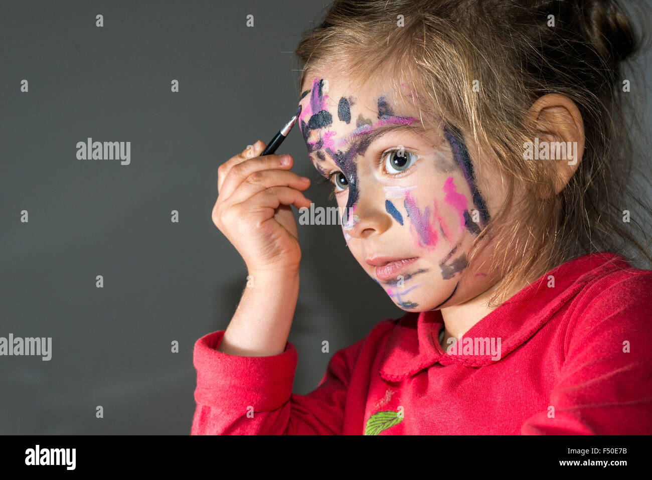 Ein kleines Mädchen, trug einen roten Pullover, ist Malen ihr Gesicht mit farbenfrohen Aquarell Stockfoto
