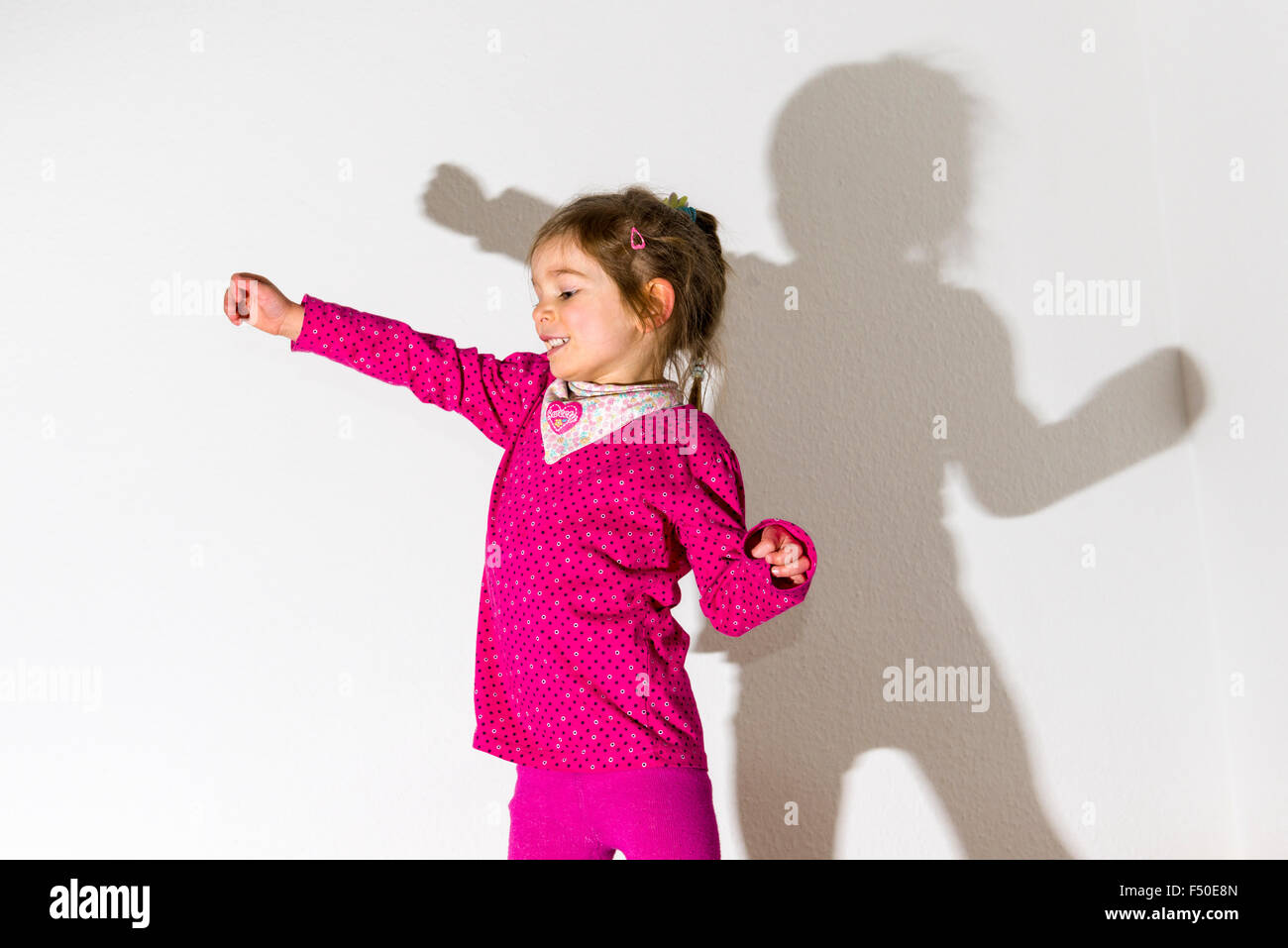 Eine blonde drei Jahre altes Mädchen, trägt ein rosafarbenes Hemd, Tanzen ist vor einer weißen Wand, ihr Schatten hinter ihr verlassen Stockfoto