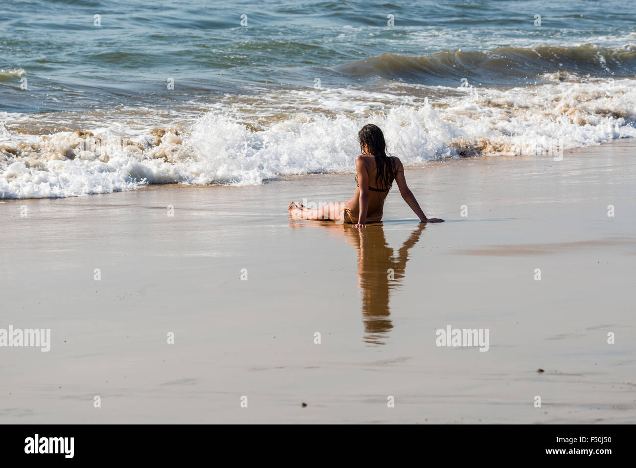 Eine junge Frau in Bikini sitzt im Sand von Anjuna Beach, mit Blick auf die kommenden Wellen Stockfoto