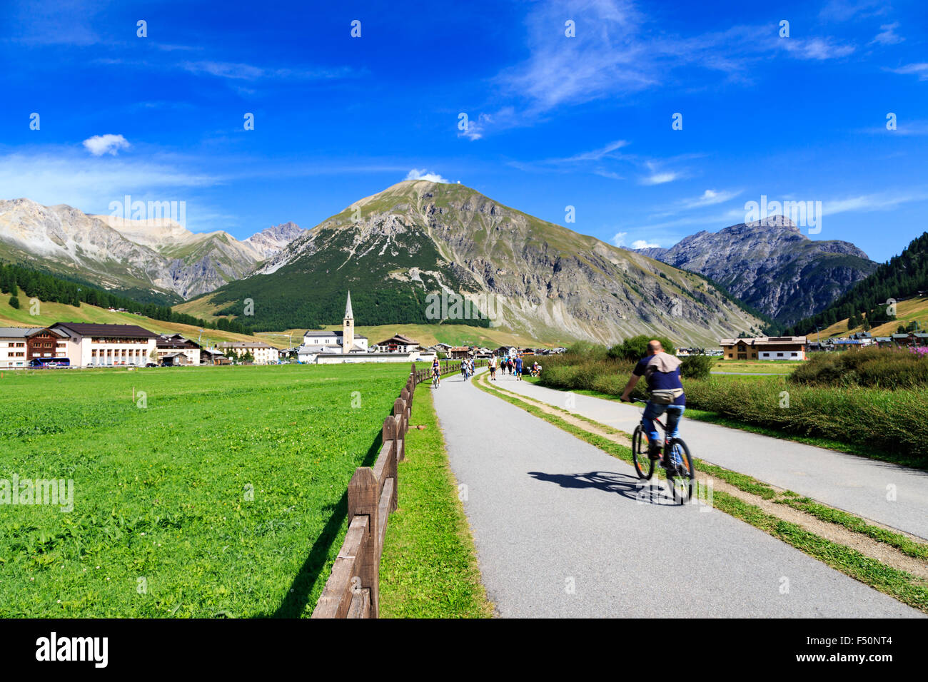 Mann Radfahren entlang einer Straße durch einen grünen Tal in Livigno, Italien in Richtung einer weit entfernten Stadt am Fuße der majestätischen Alpen Erbse Stockfoto