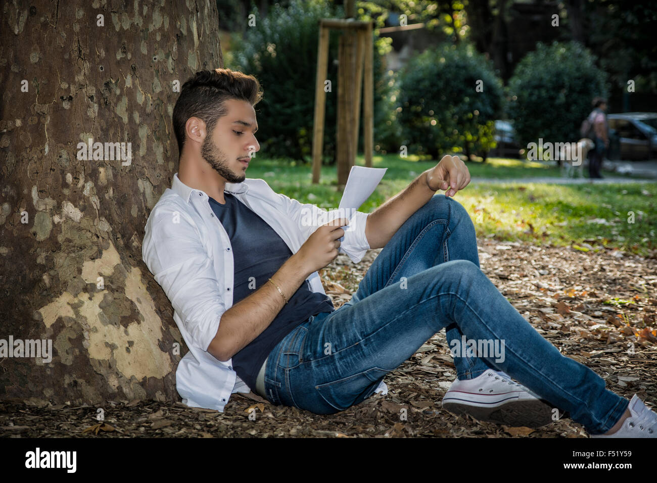 Lächelnden schönen jungen Mann, lesen oder studieren auf Papierbögen, entspannend auf grasbewachsenen Boden im Park, an Baum gelehnt Stockfoto