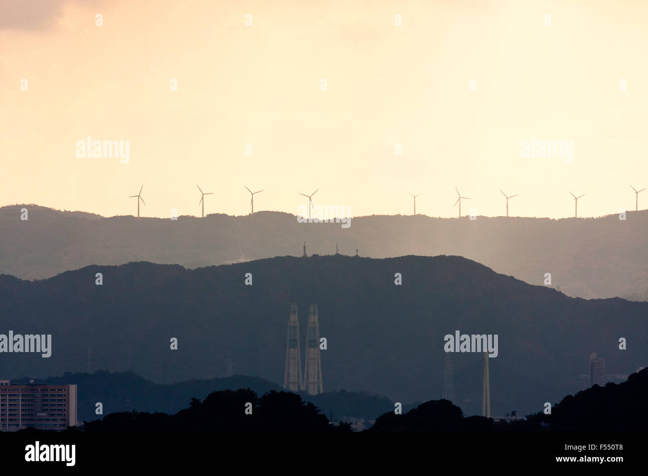 Wakayama, Japan, silhouetted Windenergieanlagen auf die Berge in der Ferne mit verschiedenen Industrieschornsteinen im Vordergrund. Regen, Sturm, aber einige Sonnenlicht. Stockfoto