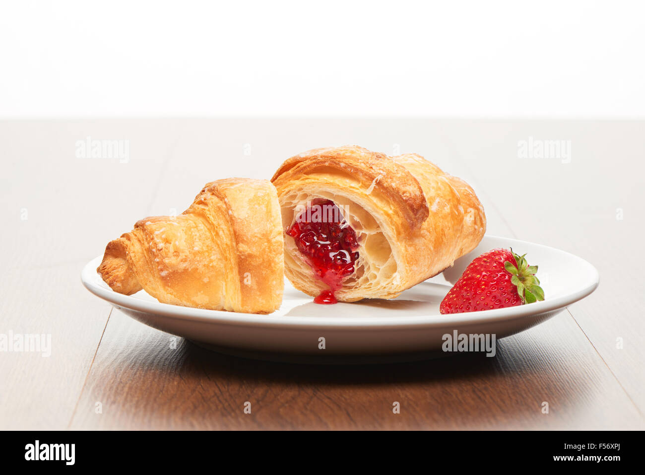 Frische französische Croissant mit Erdbeerkonfitüre ausfüllen in zwei Stücke schneiden und Erdbeere auf weißen Keramikplatte auf helles Licht bro Stockfoto