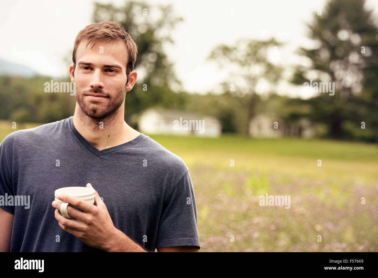 Ein junger Mann hält ein Kaffee Tasse steht in einem Feld am See. Stockfoto