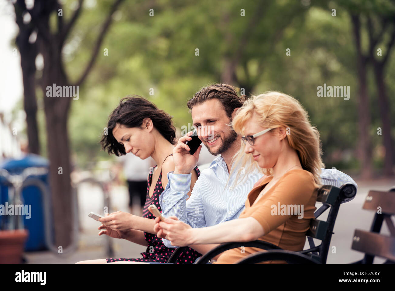 Ein Mann und zwei Frauen sitzen auf einer Bank in einem Park, überprüfen ihre Telefone, eine anrufen. Stockfoto