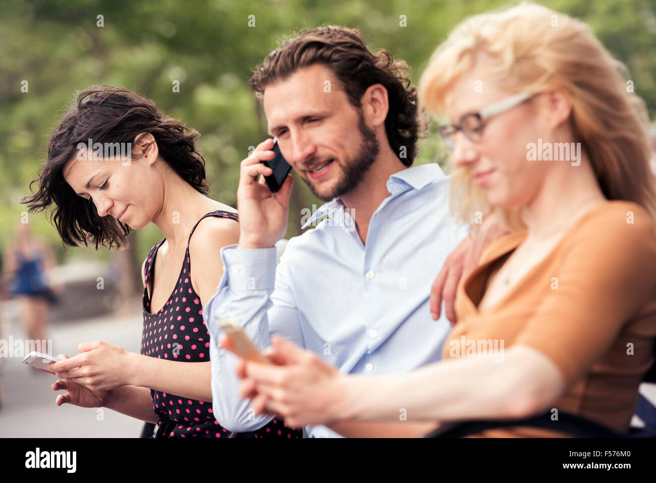 Ein Mann und zwei Frauen sitzen auf einer Bank in einem Park, überprüfen ihre Telefone, eine anrufen. Stockfoto