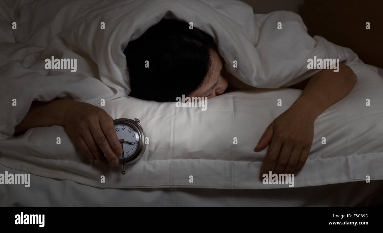 Frau mit Schlaflosigkeit, unter der Decke nach unten, halten Wecker in der hand. Wählen Sie Licht und konzentrieren Sie sich auf Frau und Uhr Stockfoto