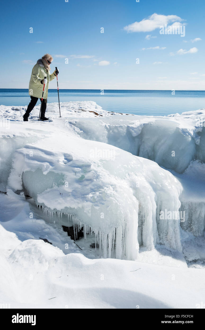 Ältere Erwachsene kaukasischen Frau herüber sorgfältig mit Skistöcke eisigen Schnee bedeckten Felsen entlang der gefrorenen Ufer des Lake Superior Stockfoto