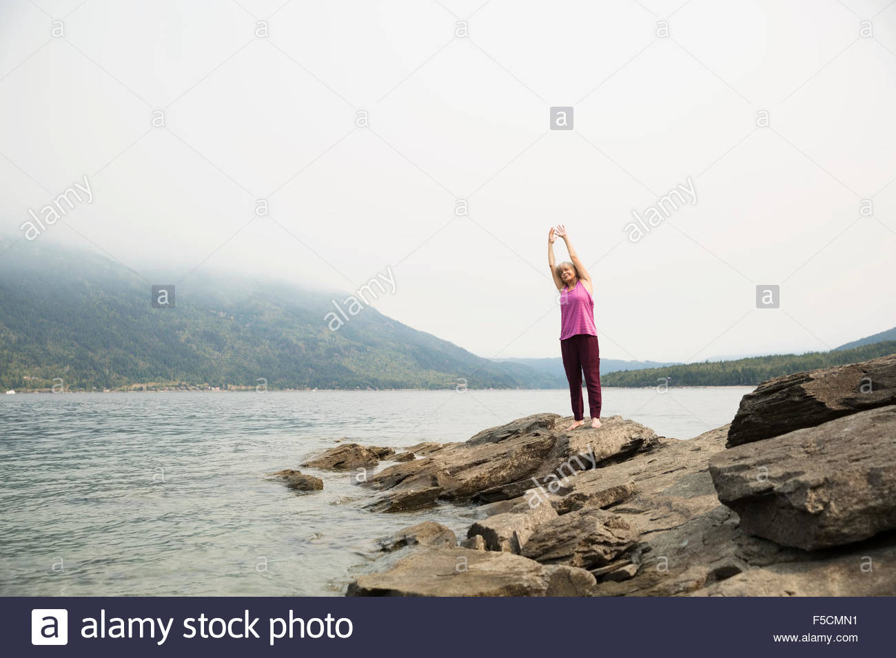 Reife Frau praktizieren Yoga auf Felsen am See Stockfoto