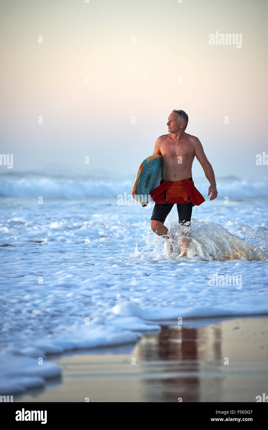 Applying Mann zu Fuß in Ozean Wellen Waschen hält ein Surfbrett. Weiche frühen Morgenlicht wäscht über die herrliche Szenerie. Stockfoto