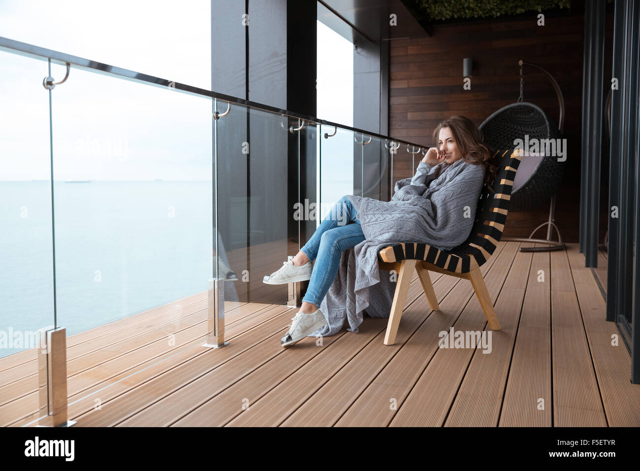 Lächelnde junge Frau sitzt in gestrickte Tagesdecke auf dem Balkon Glas gewickelt Stockfoto