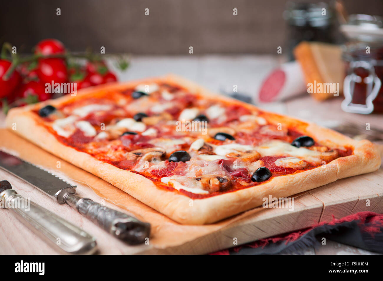 Hausgemachte rechteckige Peperoni-Pizza auf einem rustikalen Tisch mit Zutaten. Stockfoto