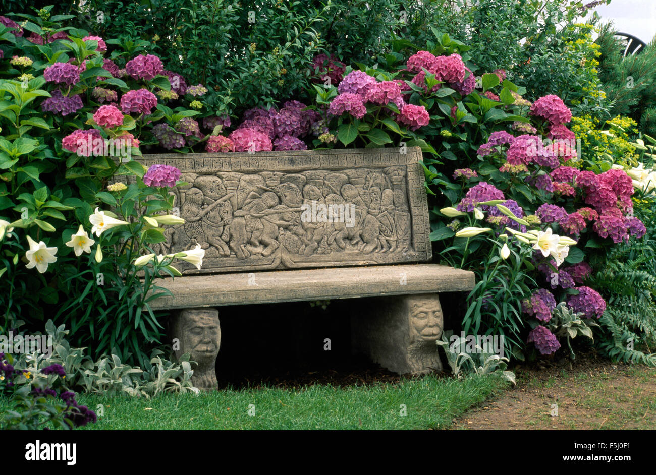 Geschnitzte Steinbank vor Grenze mit königlichen Lilien und rosa Hortensien Stockfoto