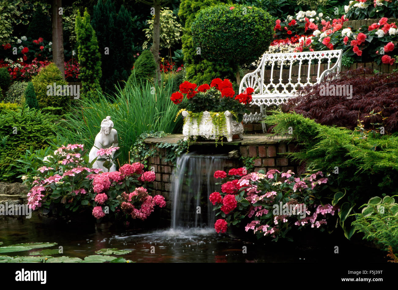 Rosa und rote Hortensien wachsen neben eine kleine Statue am Ufer neben einem kleinen Wasserfall in einen großen Garten Stockfoto