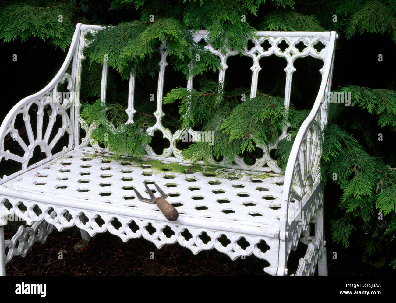 Kleinen Garten Gabel auf einem weißen Schmiedeeisen Gartenbank vor ein grüner Nadelbaum-Strauch Stockfoto
