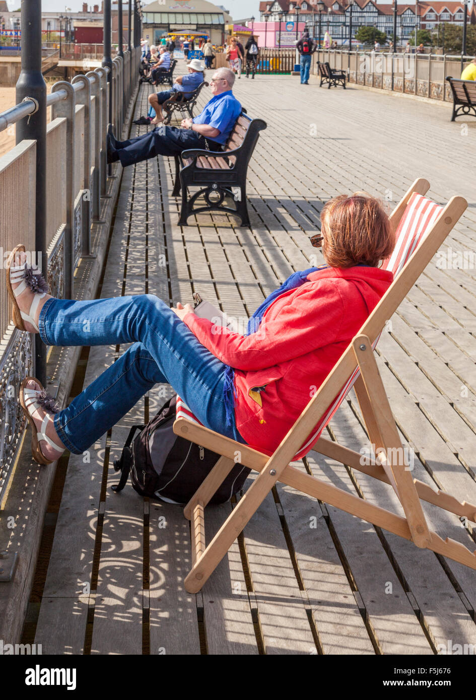 Person sitzt auf einem Liegestuhl und andere Leute saßen auf Bänken in Skegness Pier, Skegness, Lincolnshire, England, Großbritannien Stockfoto