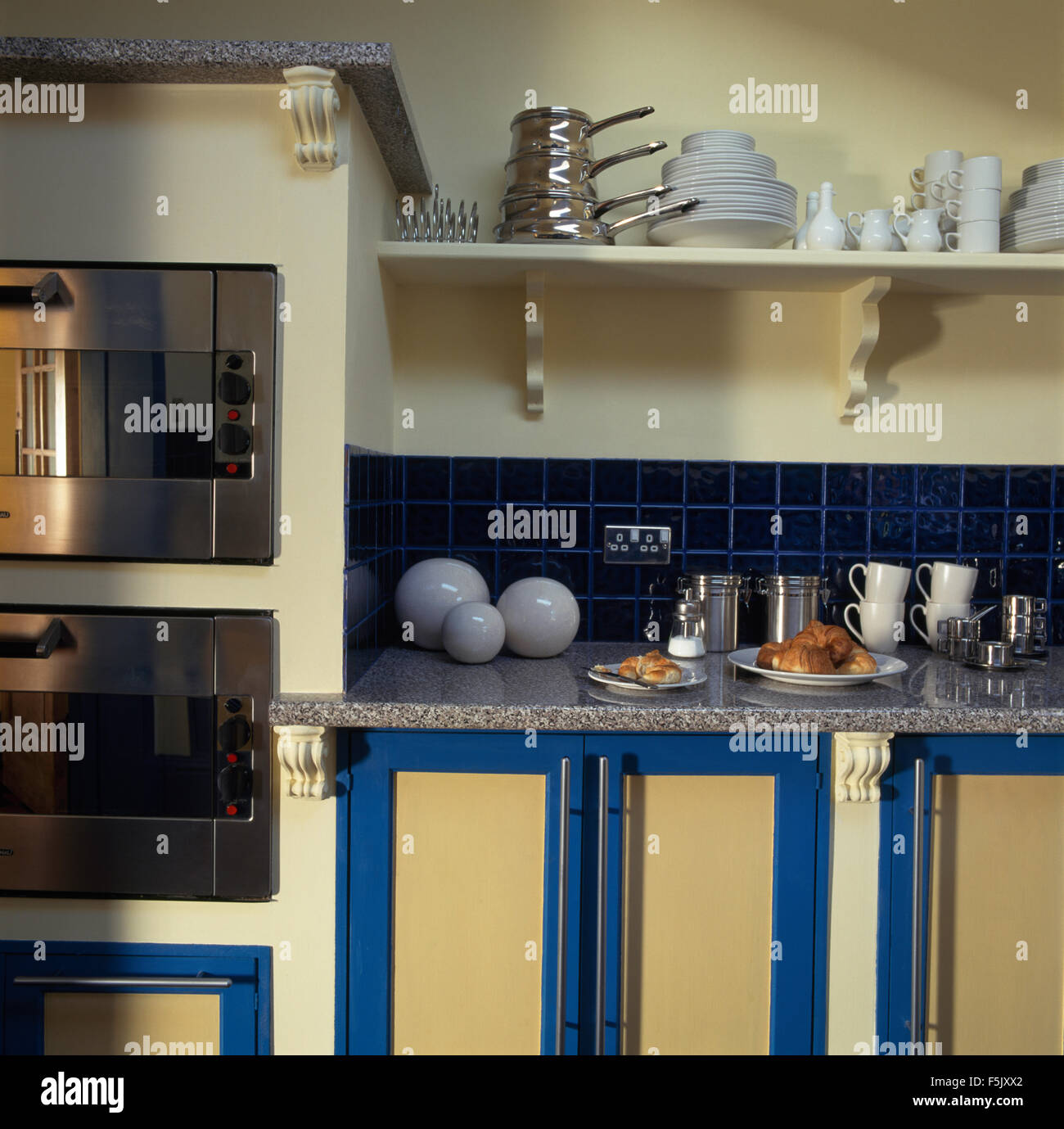 Edelstahltöpfe und weißes Geschirr auf Regal über Granit-Arbeitsplatte mit Platten aus Croissants und weiße Tassen Stockfoto