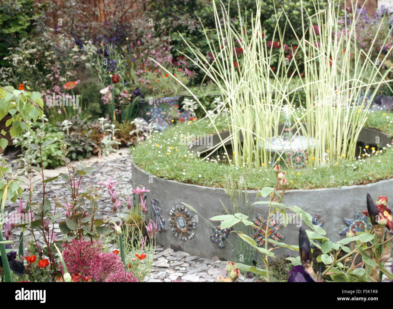 Hohe Gräser wachsen in erhöhten kreisförmigen Pool umgeben mit einem Schiefer Pfad in einen Garten im Sommer Stockfoto