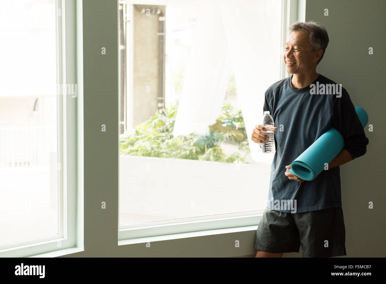 Reifer Mann hält aufgerollt Yogamatte und Flasche Wasser Blick aus Fenster Stockfoto
