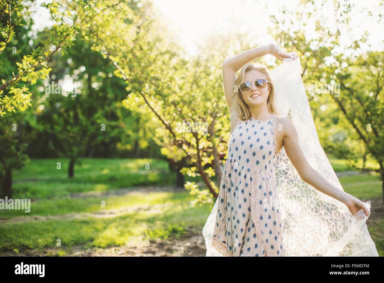 Junge Frau im Obstgarten ärmelloses Kleid und Sonnenbrille hält Spitze Stoff, Blick in die Kamera Lächeln auf den Lippen tragen Stockfoto