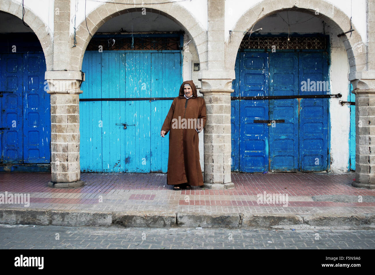 Ein Mann gekleidet in ein Djellaba lehnt sich an eine Spalte vor geschlossenen Geschäften in Essaouira, Marokko Stockfoto