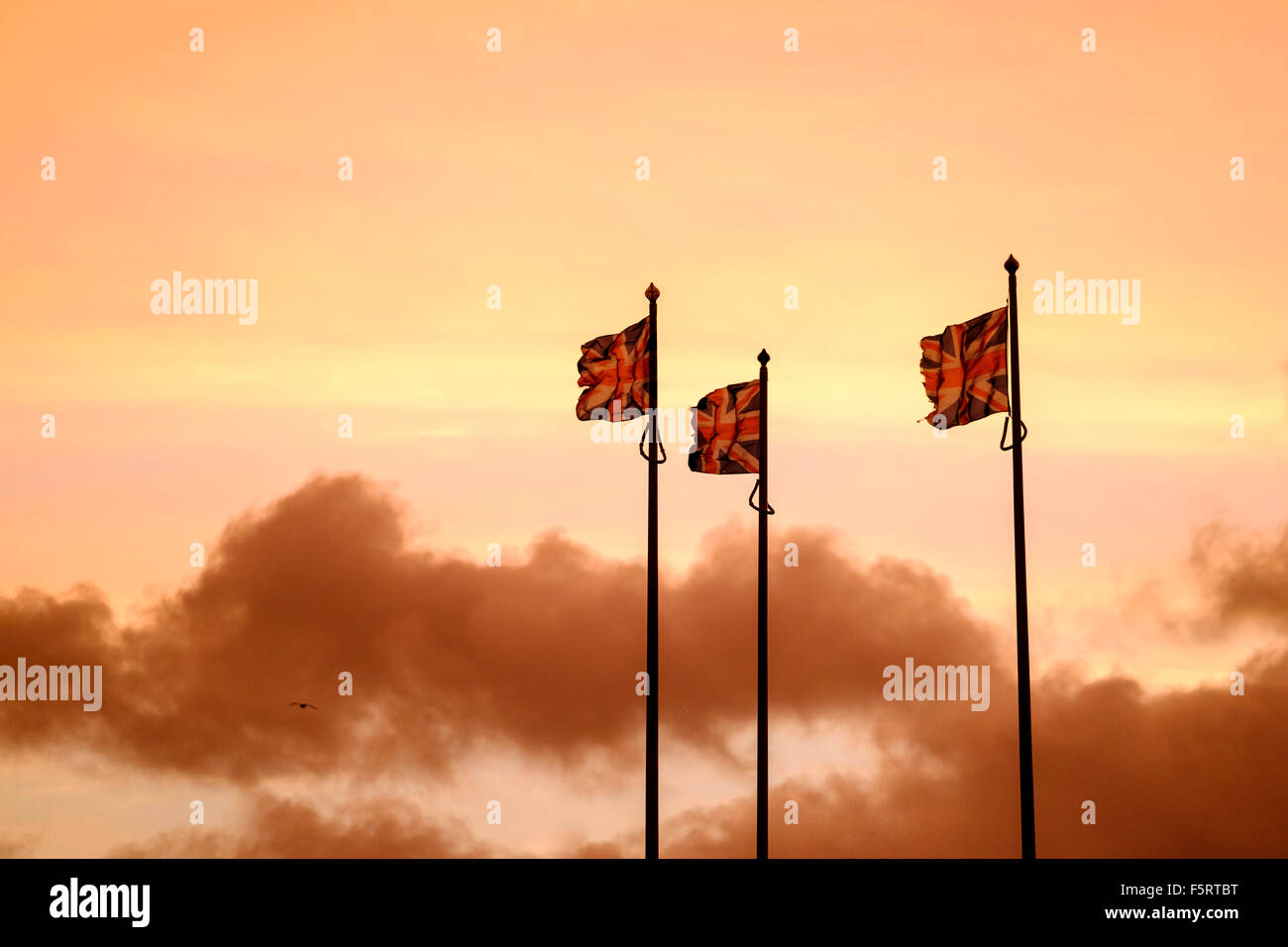 3 Union Jack-Flaggen, die bei starkem Wind gegen eine orange Sonnenaufgang Himmel fliegen Stockfoto