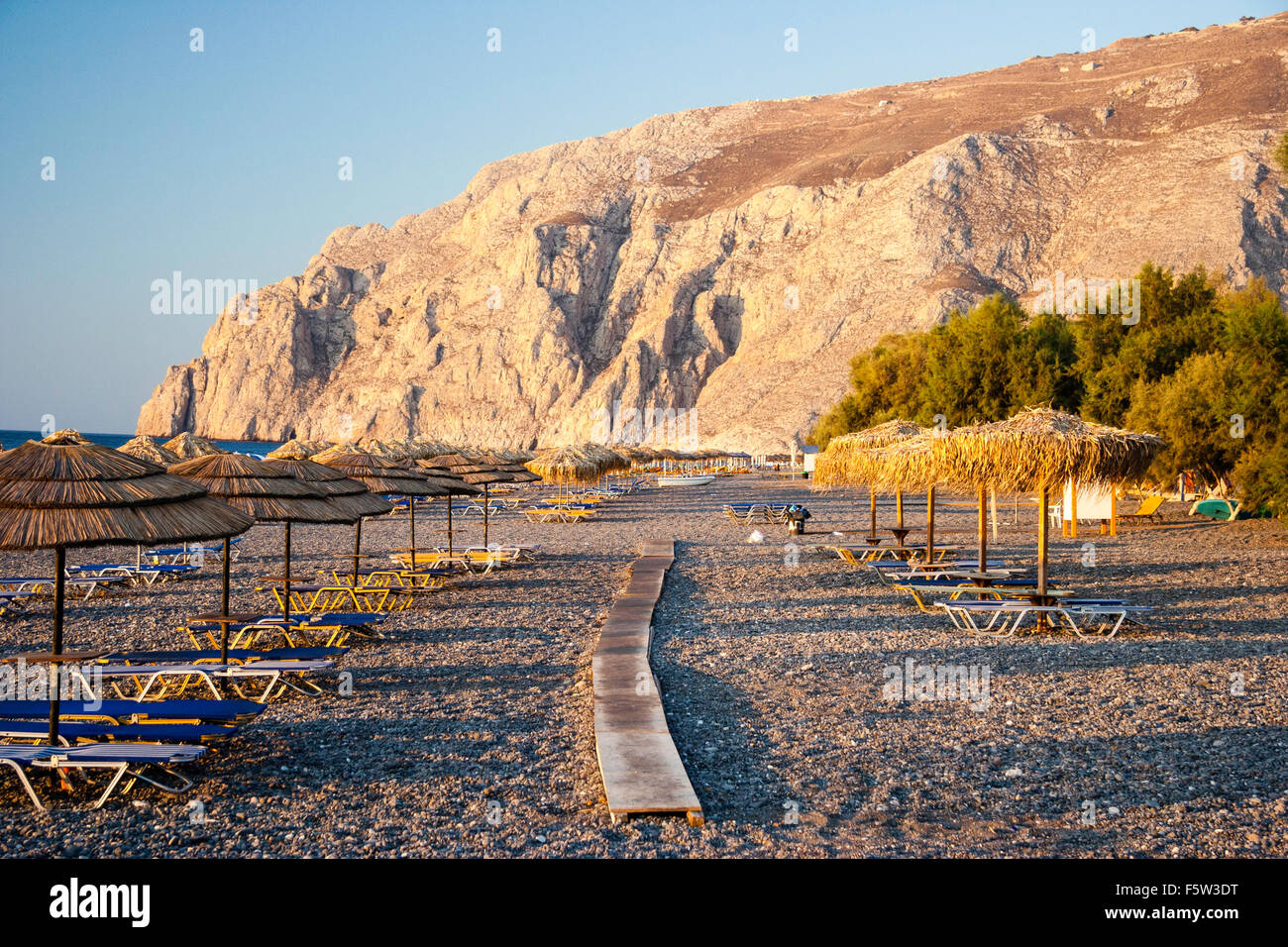 Santorini Thira. Dämmerung Blick entlang duckboards zwischen den Reihen von Sonnenschirmen und Strandliegen am leeren Strand von Kamari Resort laufen. Mase Vounco im Hintergrund. Stockfoto