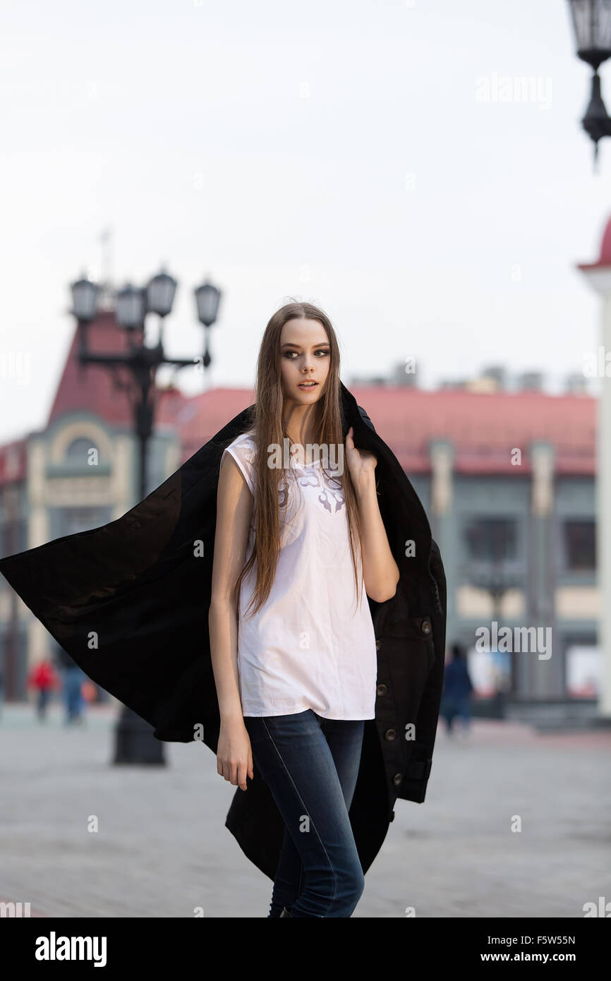schöne Mädchen geht auf dem Platz in der Stadt mit einem schwarzen Mantel Stockfoto