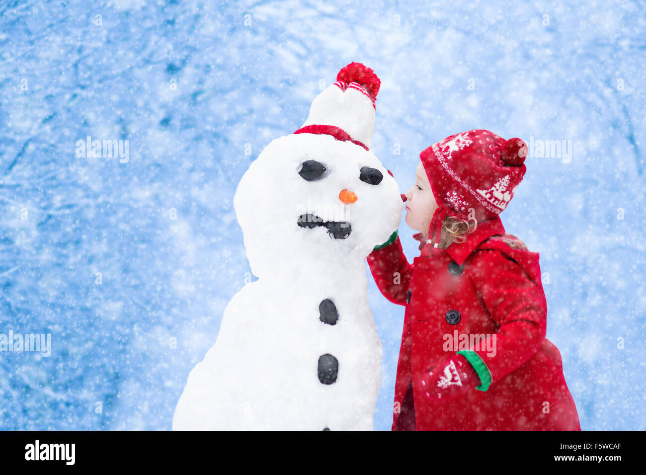 Lustige kleine Kleinkind Mädchen in einem roten gestrickte Nordic Mütze und warmen Mantel mit einem Schneemann spielen. Kinder spielen im Freien im Winter. Stockfoto