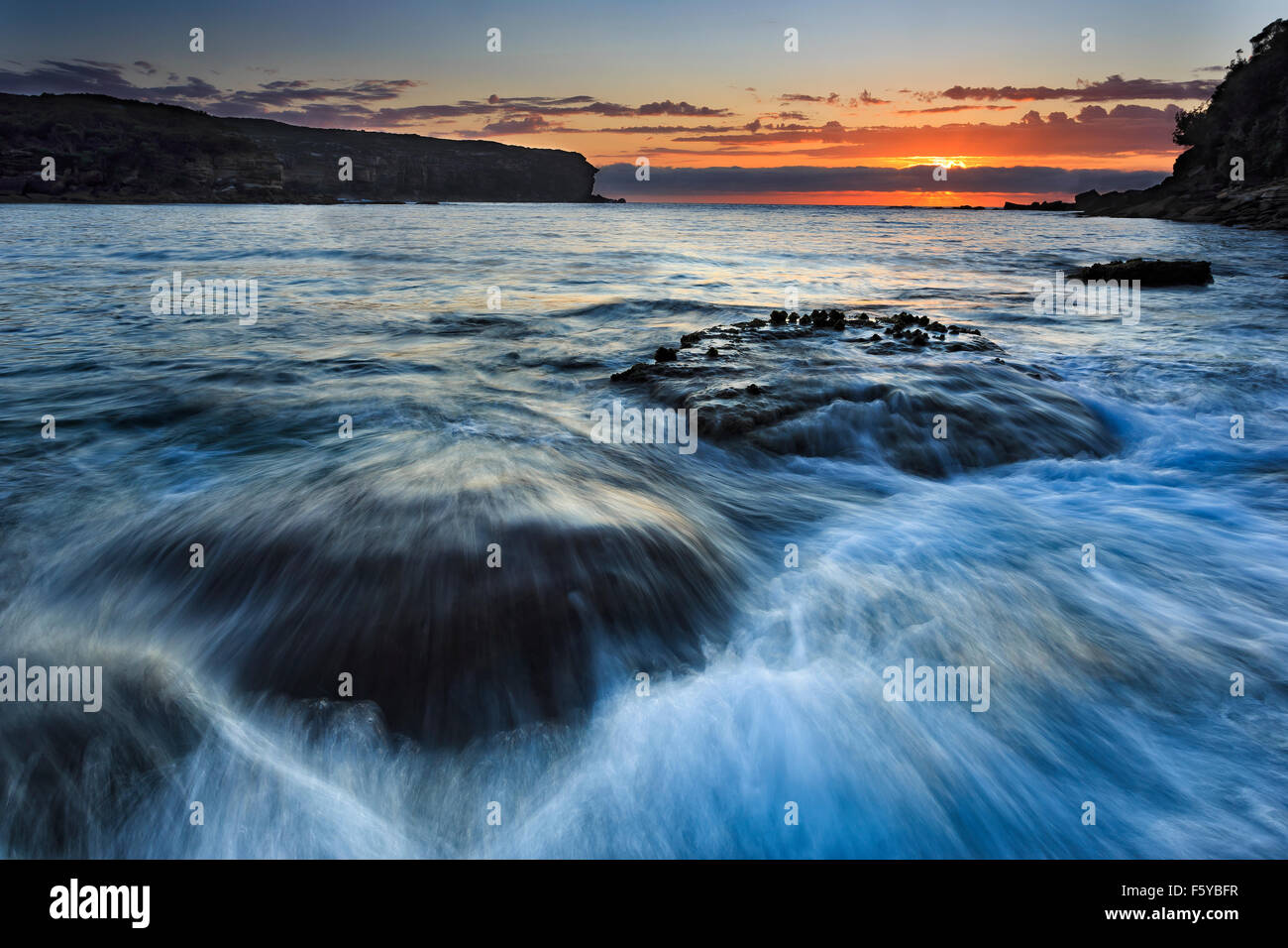 Royal National Park Wattamola Strand in Australien bei Sonnenaufgang ankommende Welle über Felsbrocken gegen eine Anhebung der Sonne über den Horizont der paci Stockfoto