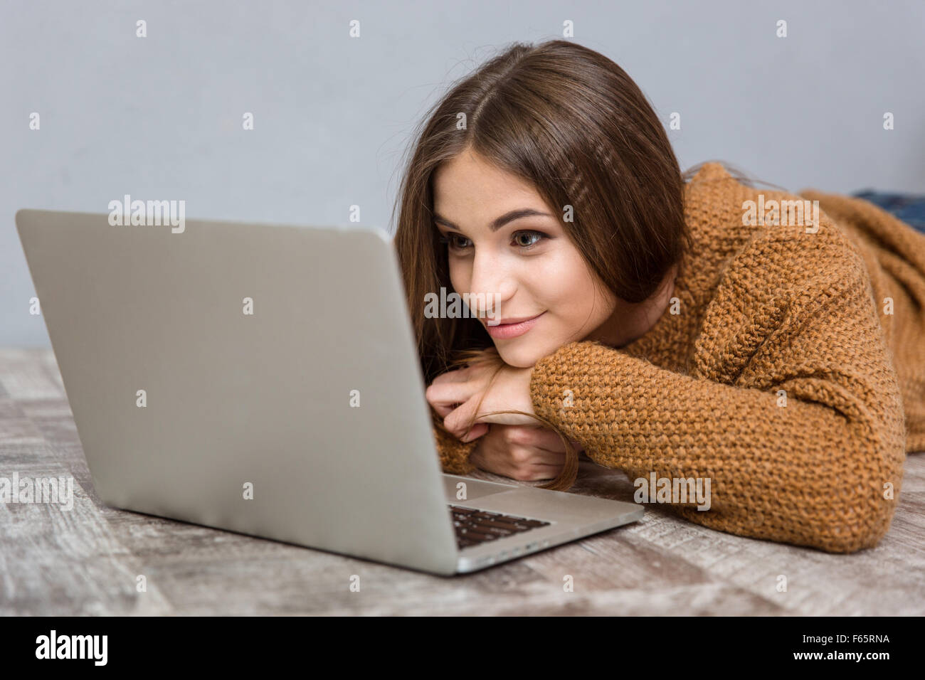 Schöne attraktive glücklich entspannt junge Frau in braunen Pullover liegend auf Holzboden mit laptop Stockfoto