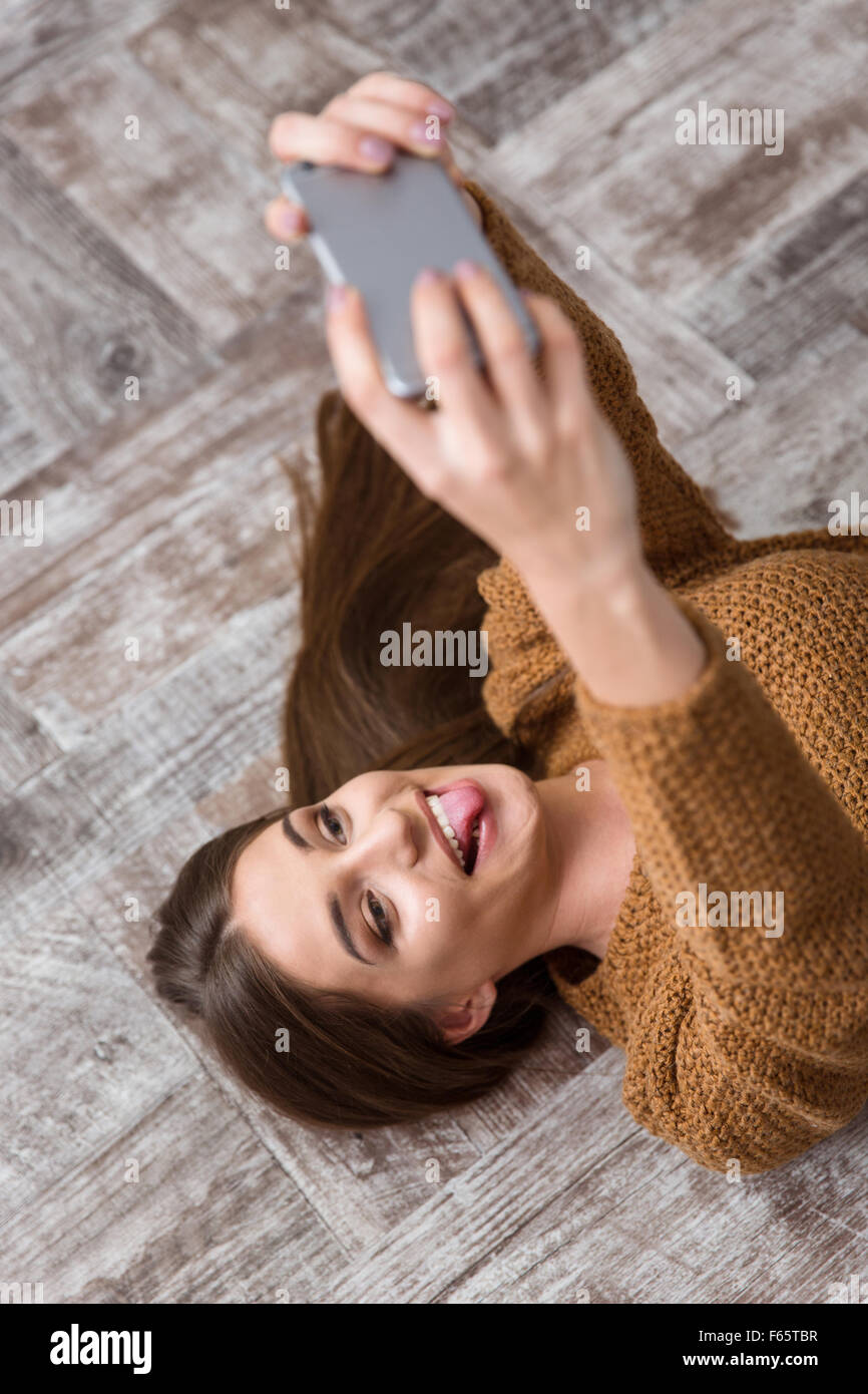Ziemlich lustige amüsante glücklich niedliche Mädchen mit langen Haaren in braunen Pullover liegend auf Holzboden, machen Selfie und Zunge zeigen Stockfoto