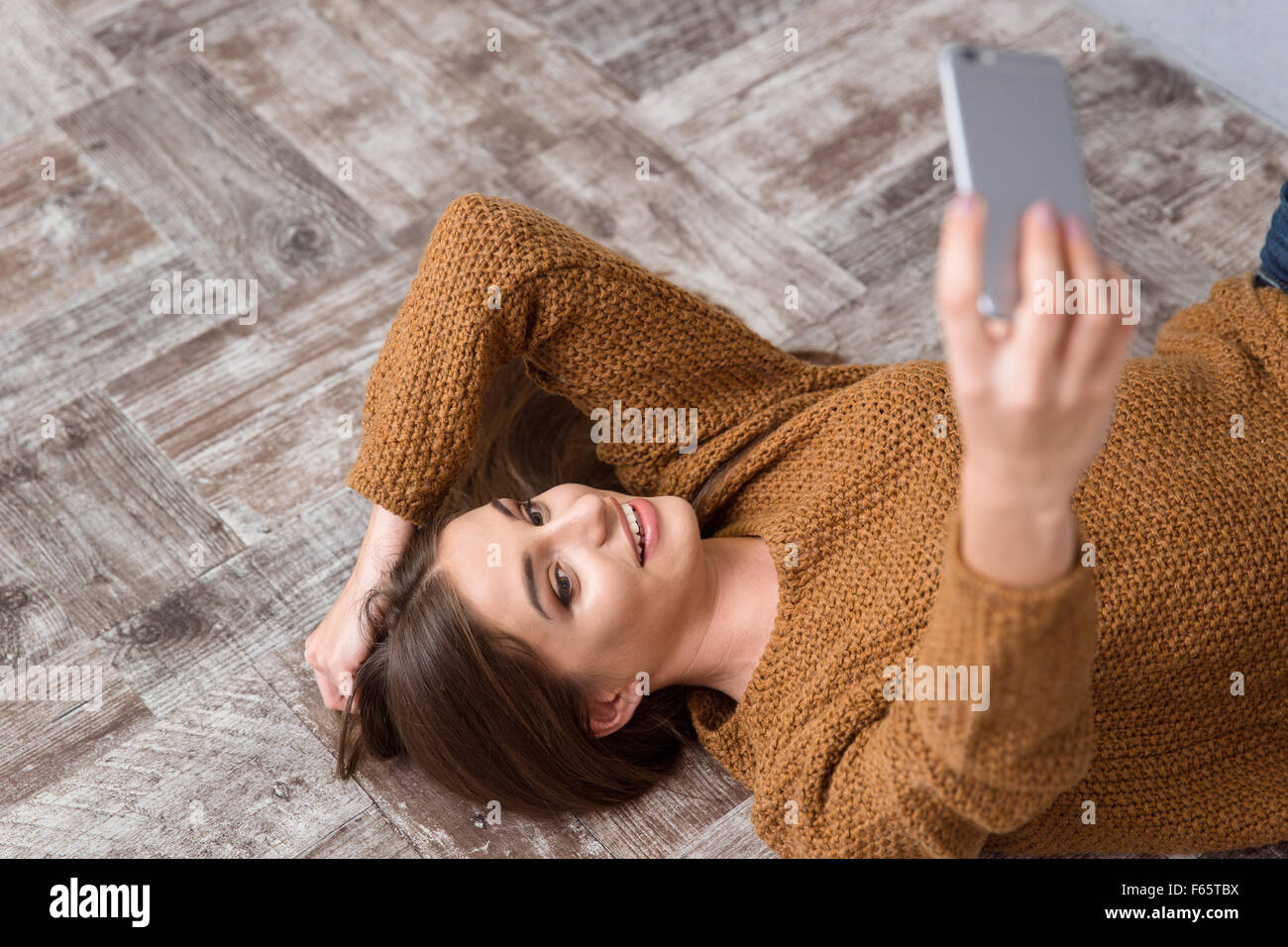 Schöne attraktive charmante lächelndes Mädchen auf hölzernen Boden liegend in braunen Pullover und Selfie zu machen Stockfoto