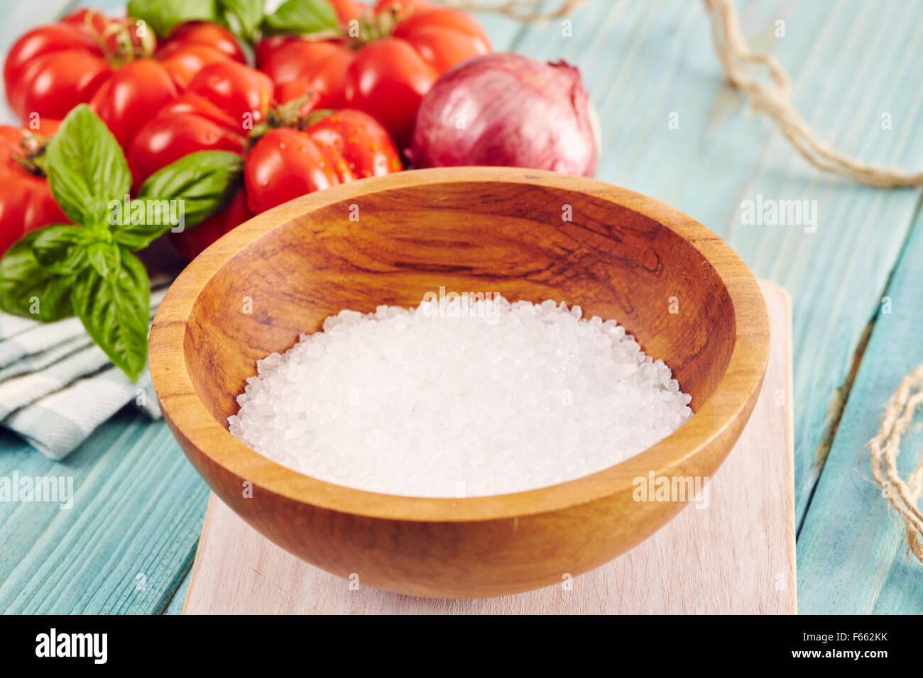 ein hölzernes Schüsselchen mit marine Salzkristalle umgeben von Tomaten und Zwiebeln auf einem Aquamarin Holztisch Stockfoto