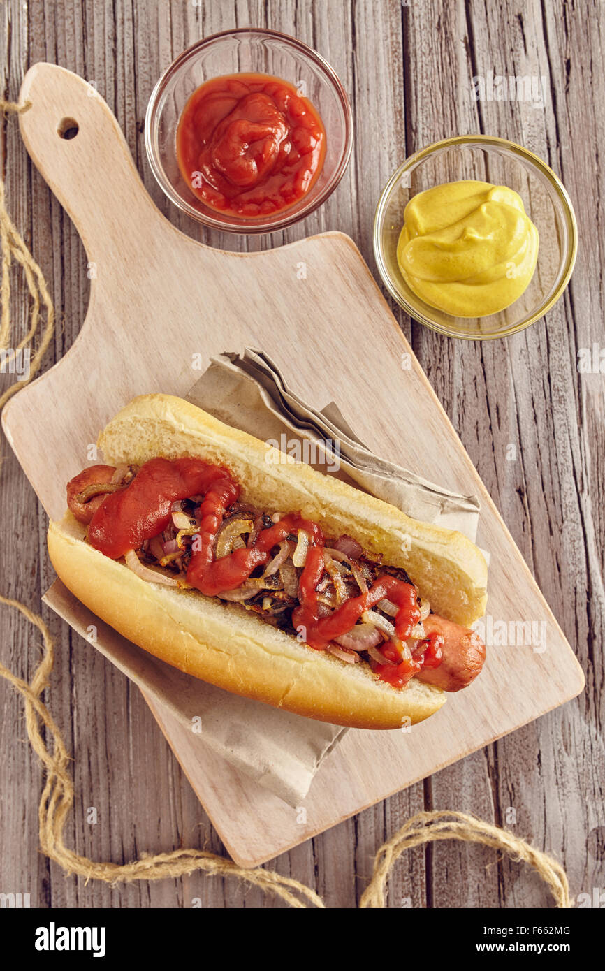 Hot Dog mit Zwiebeln, Ketchup und Senf auf einem weißen Schneidebrett auf einem Holztisch. Ketchup und Senf in zwei transparente Schüssel Stockfoto
