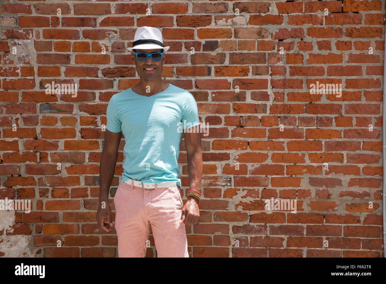 Ethnisch vieldeutigen Mann mit einem hellen Grün t-shirt, rosa Shorts, Sonnenbrillen und Fedora gegen orange-Red brick wall im Freien. Stockfoto