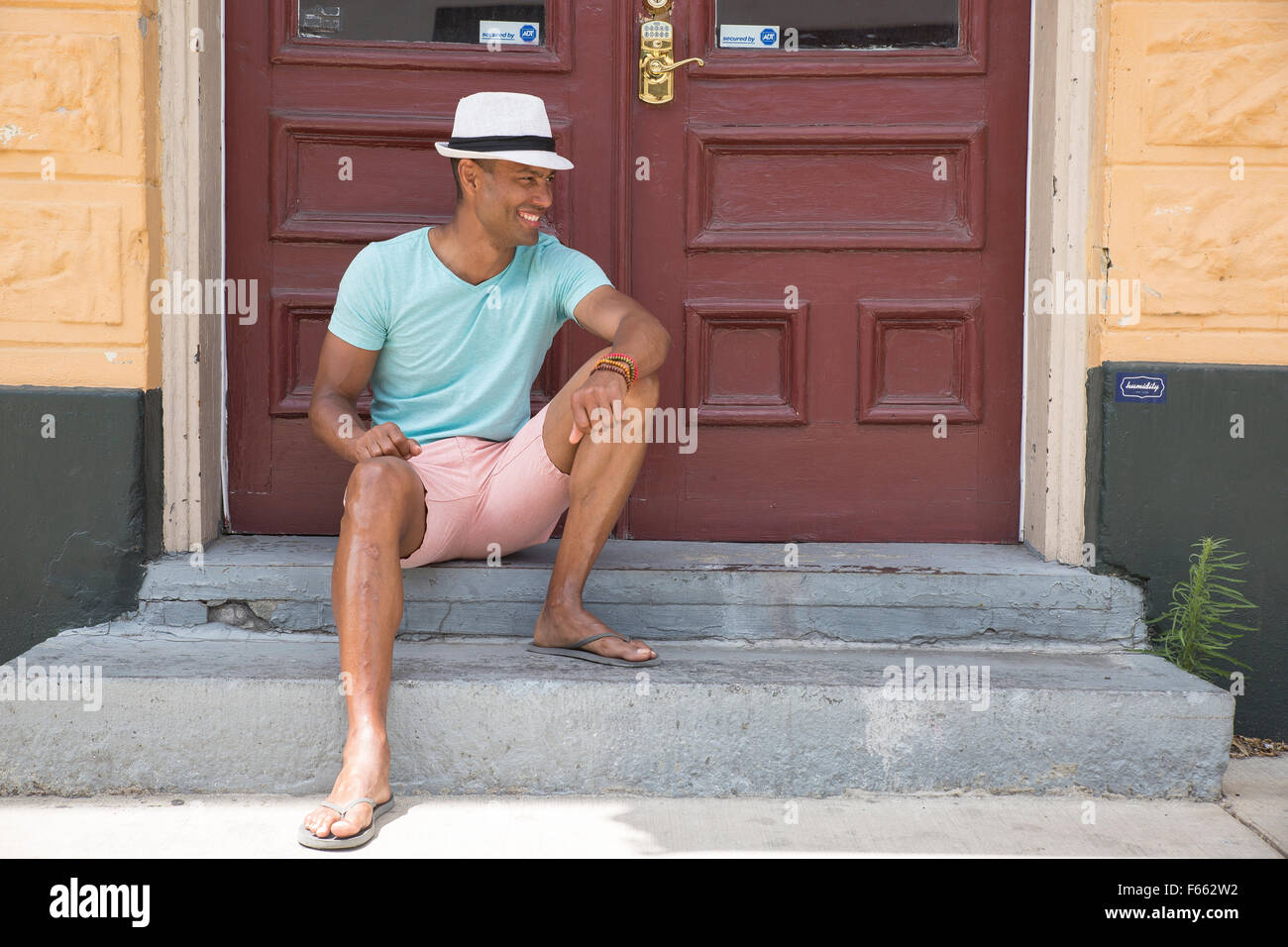Eine ethnisch vieldeutigen Mann mit einem hellen Grün t-shirt, rosa Shorts, Sonnenbrillen und Fedora auf einem Stoop außerhalb eines mit nach Hause Gebrannte Umbra Doppeltüren. Stockfoto