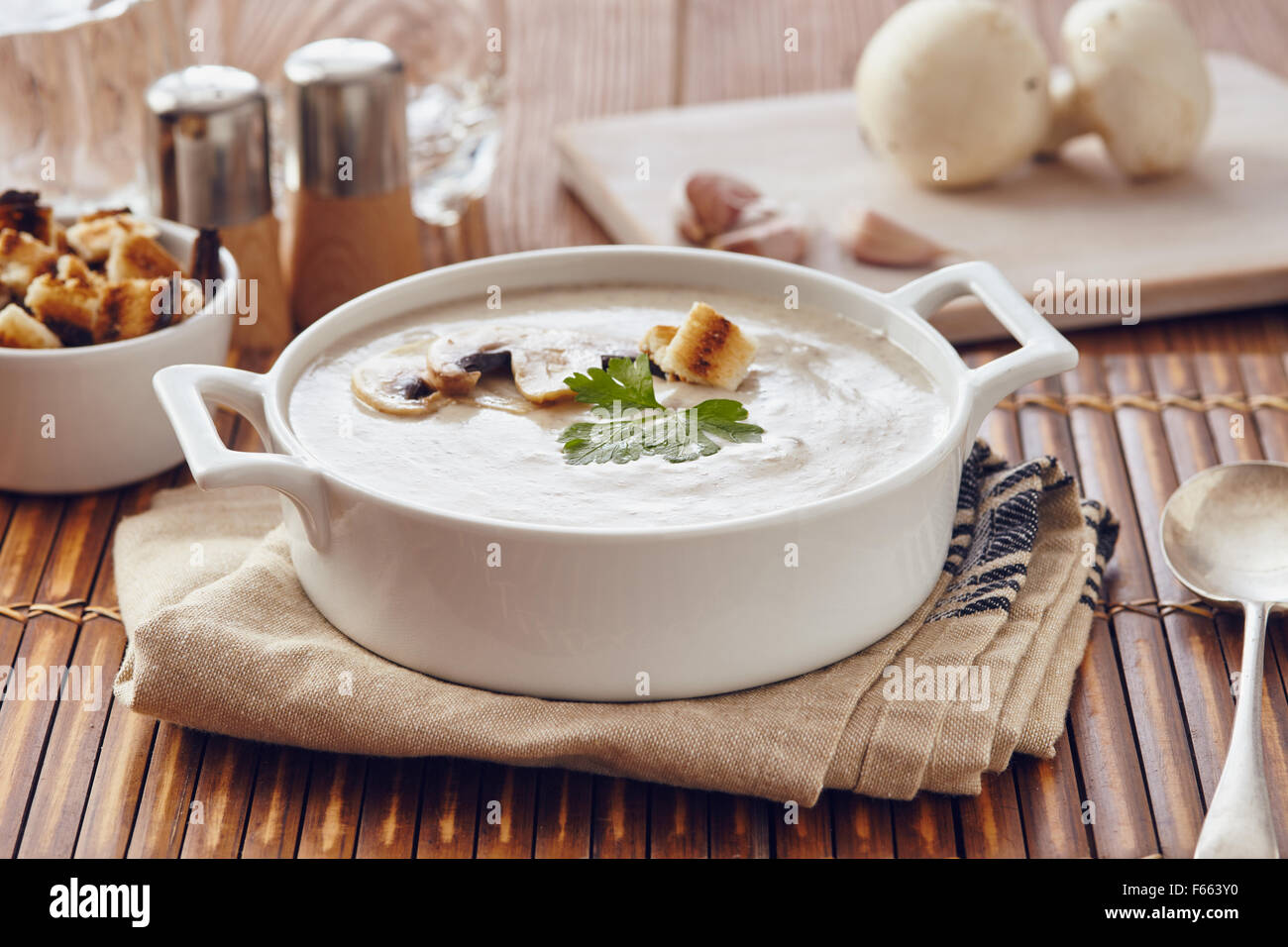 Champignon Cremesuppe auf einem Tisch mit Brot, Knoblauch und rohe Pilze auf einem Schneidebrett weiß. Salz und Pfeffer auf dem Tisch. Stockfoto